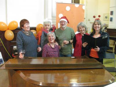 From left to right: Jane Reynolds (choir), The Green Man Gallery's Caroline Small, Fringe entries co-ordinator Ian Bowns, Joan Knox (choir) and marketing officer Stephanie Billen with choir leader Carol Bowns at the piano, all having fun at the Fringe Christmas Party 2019.